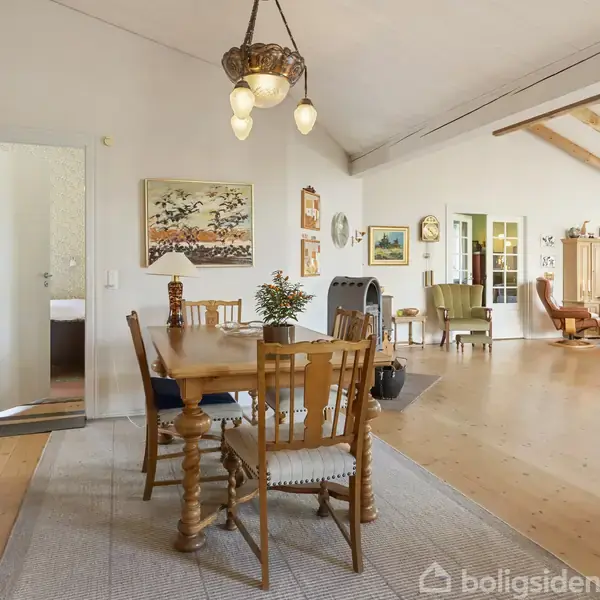 Træspisebord med seks stole står i stue med lyse vægge og trægulv. Malerier og lamper hænger på væggene, mens lænestole og skabsenheder fylder rummet. En åben dør viser et soveværelse.