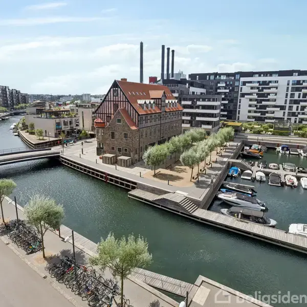 En brostensbygning med rødt tag står ved en havn, omgivet af moderne lejlighedsbygninger og båder i vandet. Kanaler strækker sig gennem byområdet med træer langs kanterne.