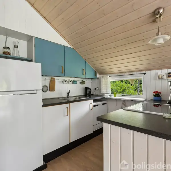 Et kompakt køkken med en hvid og blå farvepalet. Køleskab, ovn og vask er indbygget. Der er et ovenlys, og træpaneler pryder skråt loft og vægge.