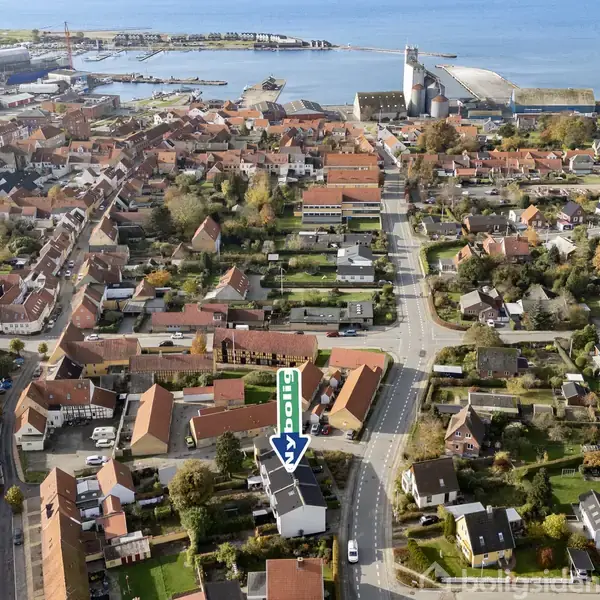 Byhus med have markeret med en blå pil i et tæt beboelsesområde nær kysten. Omkringliggende bygninger og veje strækker sig mod en havn i baggrunden.