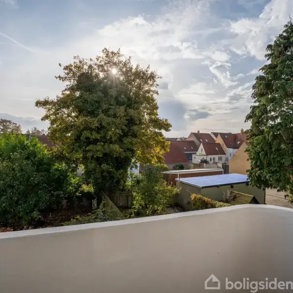 En balkon overser en have med træer, mens solen stråler gennem bladene. I baggrunden ses et boligområde med røde tage under en delvis skyet himmel.