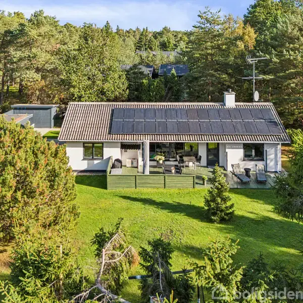 Hus med solpaneler på taget, beliggende i en grøn have omgivet af træer. En terrasse foran huset har udendørs møbler. Skovklædt område i baggrunden.