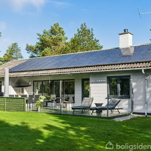 Et moderne, solcelledækket hus står stille i en grøn have omkranset af høje træer. Der er liggestole og et spisebord på terrassen, hvilket skaber en afslappet udendørs stemning.
