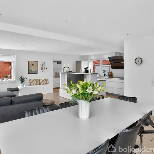 Spisestuebord med vase og blomster står i et lyst åbent rum med grå sofaer og et moderne køkken. En trappe ses til højre, og store vinduer bringer lys ind.