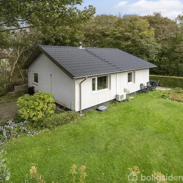 En hvid bungalow ligger på en velholdt græsplæne, omgivet af blomster og buske. I baggrunden ses en tæt skov, der danner en naturlig ramme om huset.