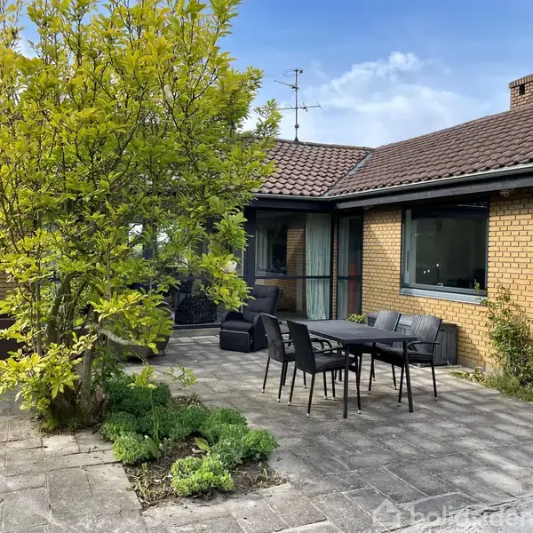 En terrasse med et sort udendørs spisebord og stole omgivet af en bungalow i gule mursten. I forgrunden står et frodigt træ, og omgivelserne er rolige og grønne.