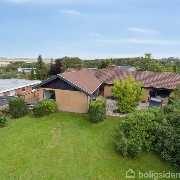 Et hus med rødt tegltag står på en velholdt græsplæne omkranset af træer og buske. Baggrunden viser et landligt område med åbne marker og skovklædte områder.