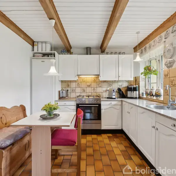 Stue med træbjælker i loftet, lædersofaer og lænestole, samt spisestue med seks stole. Røde orientalske tæpper på gulvet. Væggene har kunstmalerier og der er store vinduer med planter.