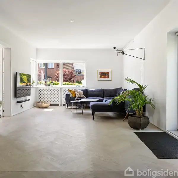 Stue med sort sofa, lamper og grønne planter i et lyst rum. Store vinduer og døre åbner ud mod haven, og en fladskærm hænger på væggen.