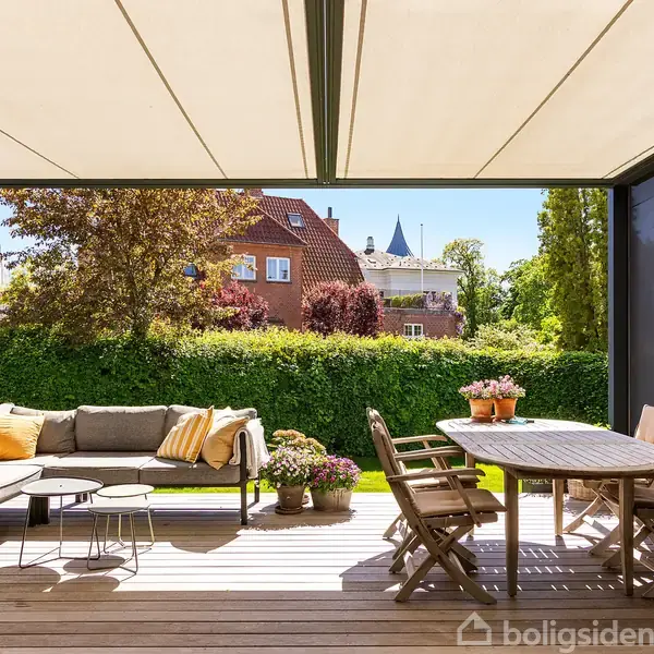 Terrassemøbler står under en åben pergola med en sofa og et rundt bord med stole. Omgivet af grønne planter med udsigt til et rødt murstenshus og klar blå himmel.