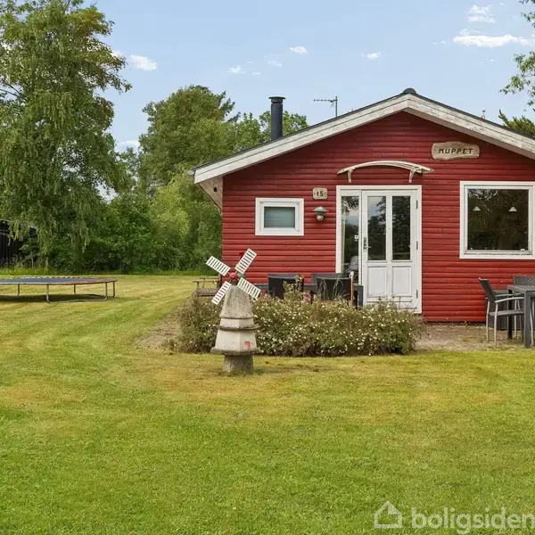 Et rødt træhus står på en græsplæne med en miniature vindmølle foran. Bord og stole står rundt om huset. Skiltet over døren lyder: "MØRFJÆRT." Trampolin og træer i baggrunden.