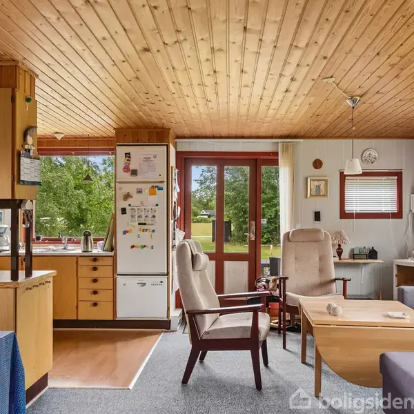 Stue med træloft har en sofa, lænestole og et sofabord. Et køkkenhjørne med køleskab og skabe er til venstre. Store vinduer og en dør viser en grøn have udenfor.
