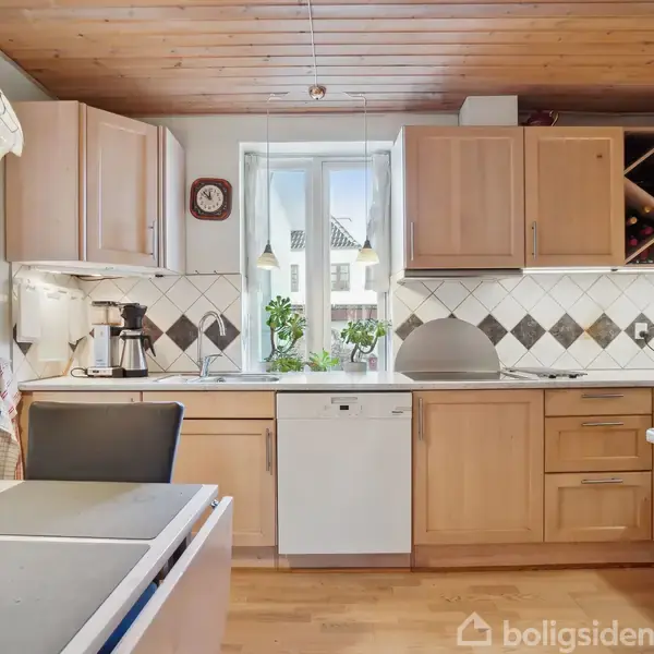 Køkken med lyse træskabe; kaffemaskine og planter på bordplade; mosaikfliser i baggrunden; spisebord i forgrunden; loft af træ; udsigt til have gennem vindue.