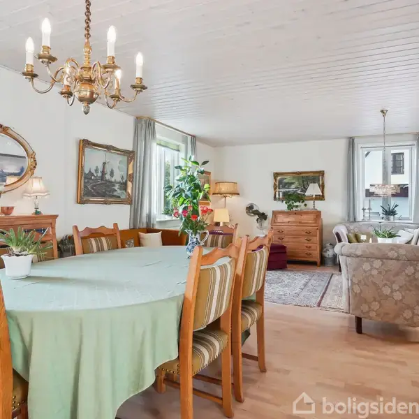 Et spisebord med en grøn dug er omgivet af stole i en stue med lyse trægulve. En lysekrone hænger over bordet, og der er planter, malerier og en sofa i baggrunden.