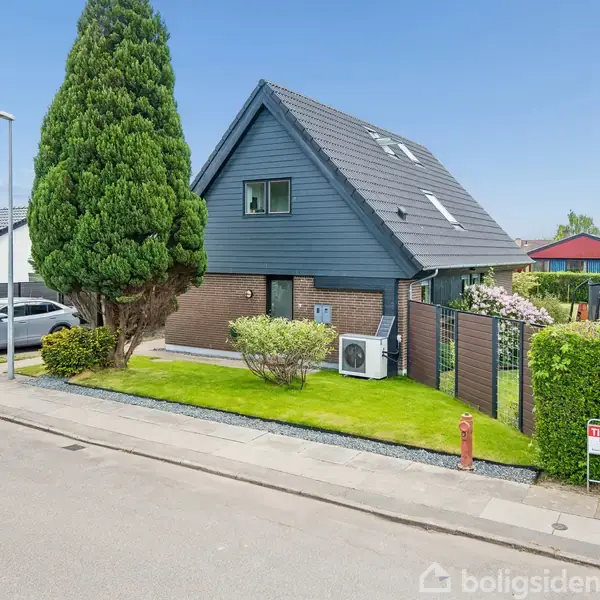 Et moderne hus med skråt tag står på en velholdt græsplæne omgivet af buske og en stor træ, beliggende ved en stille gade. Et "Til salg" skilt er placeret foran.