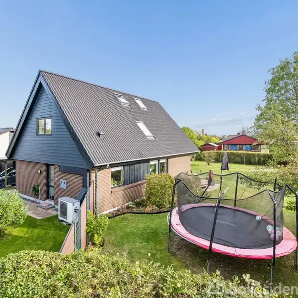Hus med spidstag står i en velholdt have, omgivet af buske og træer. En stor rund trampolin er placeret i græsplænen tæt ved huset.