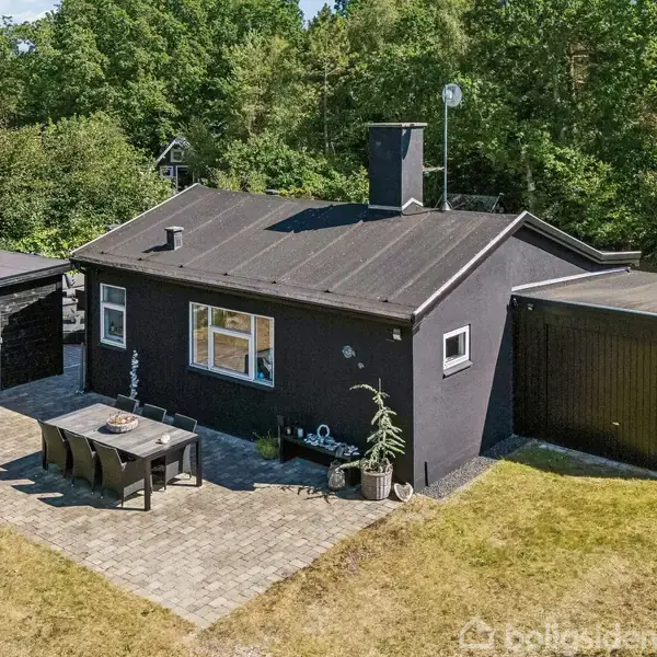 Et sortmalet hus står stille på en græsplæne med haveborde, omgivet af tæt bevoksning og skov. Ved siden af huset ses et lille skur.