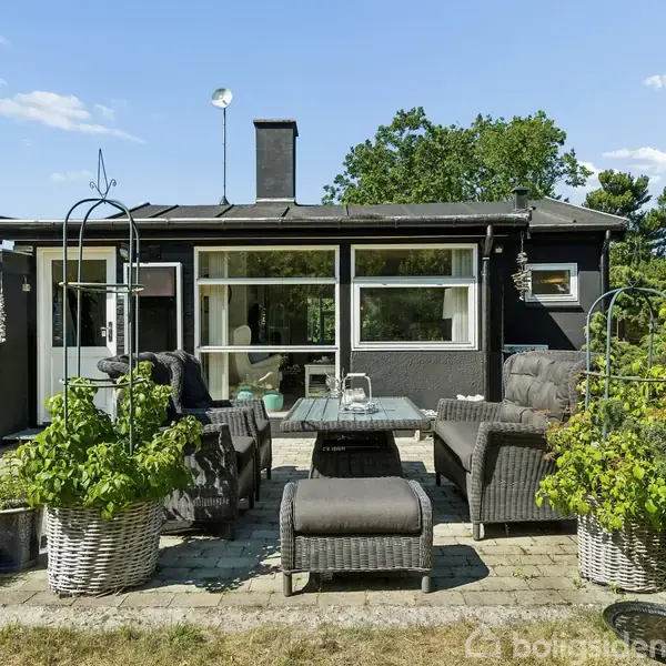 Hus med fladt tag og store vinduer står bag en terrasse. Terrasse har flettede havemøbler og planter i kurve. Omgivet af grønne buske og en klar, blå himmel.
