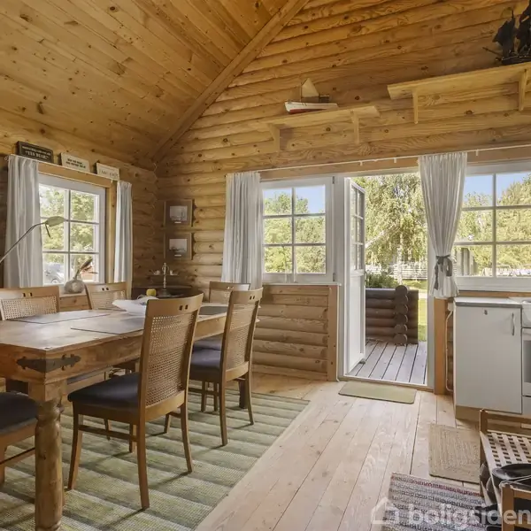 Træbord med stole står i en træhytte. Døre åbne til en terrasse. Køkkenhjørne med komfur og køkkenvask. Lysindfald gennem vinduer. Hylder og en lille dekorativ båd over døren.