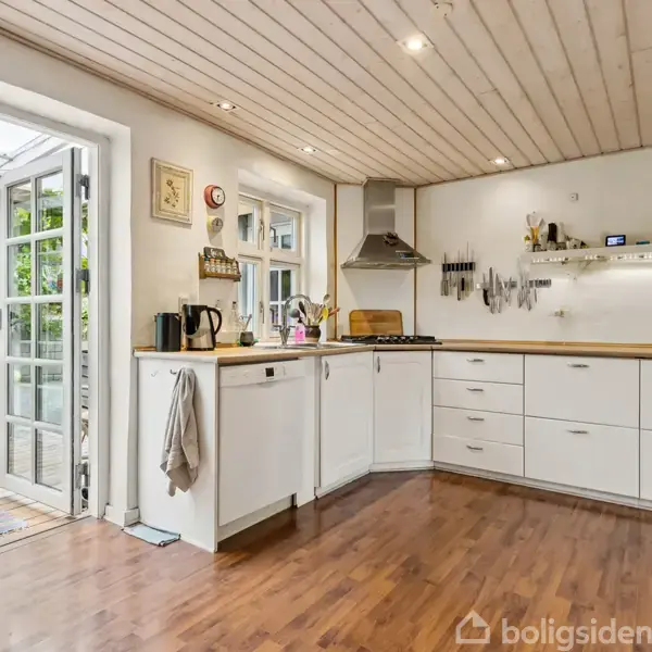 Et funktionelt køkken med hvide skabe og træbordplader. En dør står åben mod en terrasse med udsigt til en frodig have. Køkkenet har trægulv og loftspots.