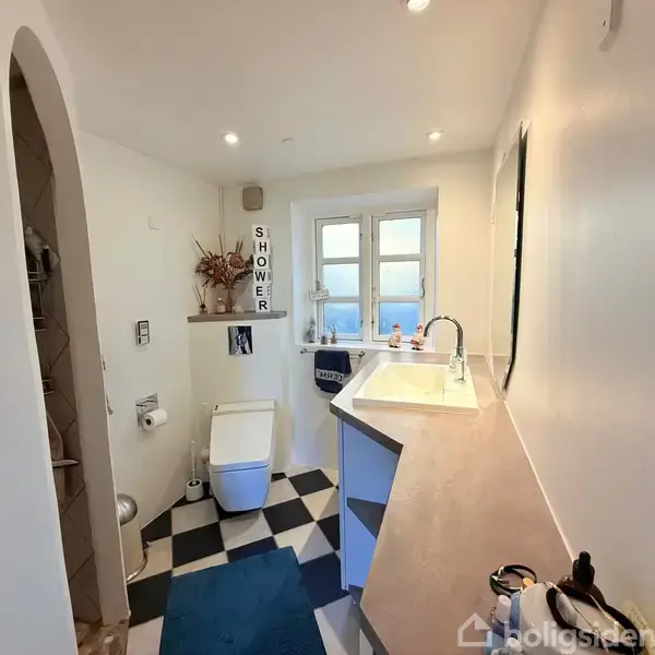 Et lyst badeværelse med toilet og vask, hvor et skilt med teksten "SHOWER" hænger ved et lille vindue. Sort-hvide fliser dækker gulvet.