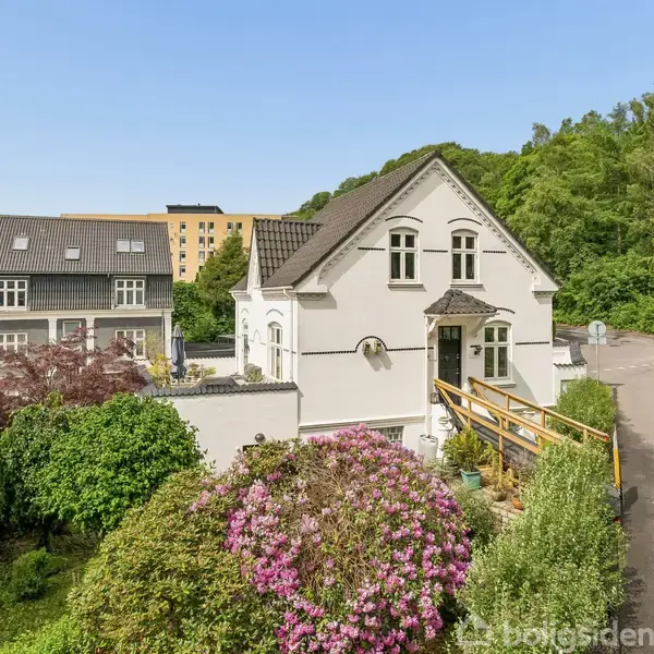 Et hvidt hus står i fokus, omgivet af en frodig have med blomstrende buske. Til venstre ses et nabohus, og i baggrunden er der skov og en lys himmel.