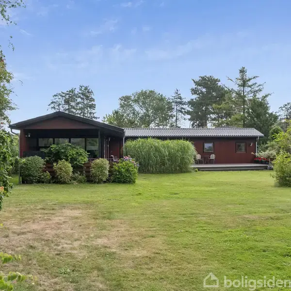 Et rødt træhus med store vinduer står i en velplejet have. Omkring huset er der tætte buske og høje træer under en klar blå himmel.