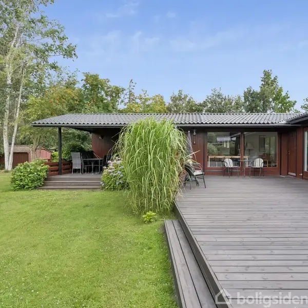 Hus med stor træterrasse omgivet af græsplæne og træer. Terrassen har udendørs siddepladser og planter. Huset har store vinduer og er indlejret i en grøn have.