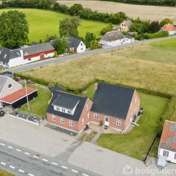 Flere huse ligger langs en vej i et landligt område. Hovedfokus er et rødt murstenshus med sort tag, omgivet af græs og marker i baggrunden.