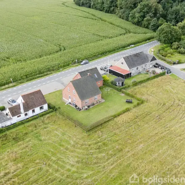 En række huse ligger langs en vej omgivet af marker, med en markant rød bygning i fokus. Baggrund byder på skovklædt område.