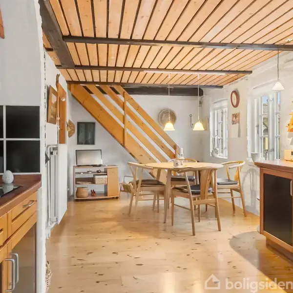 Et lyst køkken med træloft og gulv viser en spisekrog med træbord og stole. En trappe fører til en anden etage, og køkkenbordet har moderne apparater. Veggene er hvide.