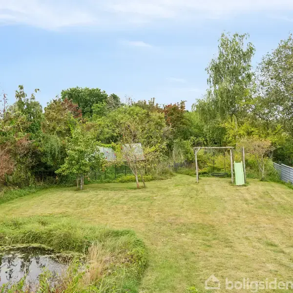 En græsplæne med en gynge står stille i en rummelig have, omgivet af høje buske og træer. En lille dam ses i forgrunden under en klar blå himmel.