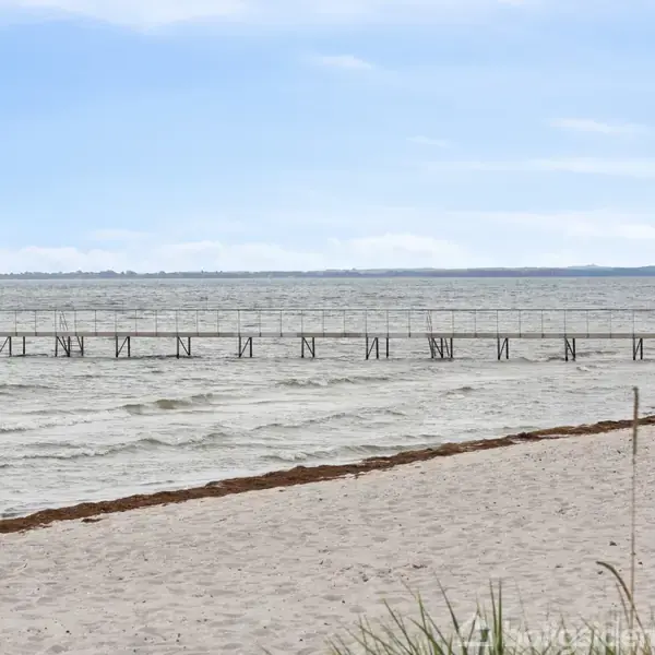 En lang badebro strækker sig ud i det rolige havvand, omgivet af en sandstrand med spredte græsstrå og en klar, blå himmel.