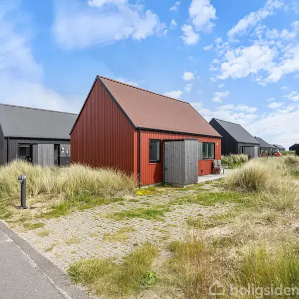 Et rødt træhus står stille blandt høje klitter med flere ens huse i baggrunden. Himmelen er blå med lette skyer, og en asfalteret vej løber langs huset.
