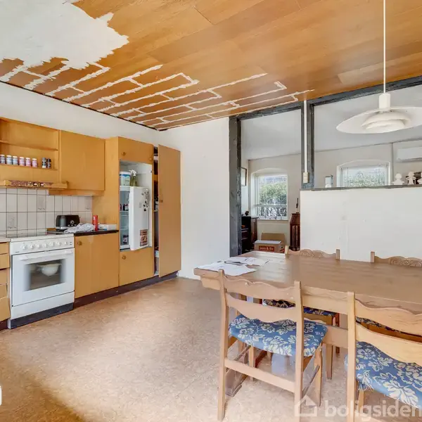 Et køkken med træskabe, hvid ovn og køleskab til venstre. Loftet har afskallet maling. Et spisebord med blå-stole står centralt. Rummets væg er delvist glas.