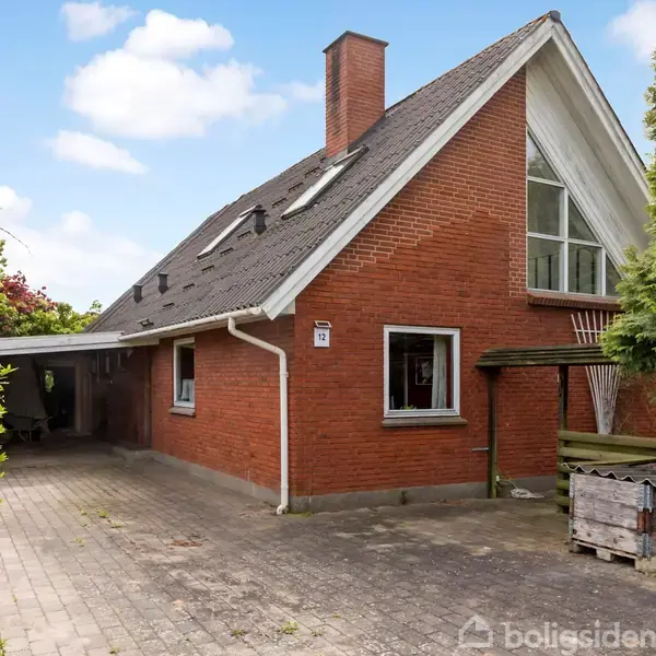 Et rødt murstenshus med sort skråt tag har en carport og er omgivet af grønne træer. Vinduerne i taget har ovenlys. En affaldsbeholder står nær indkørslen.