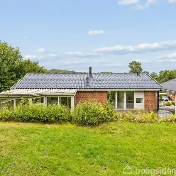 Et rødt murstenshus med sort tag står stille i en grøn have omgivet af træer. En veranda er til venstre, og en garage til højre.