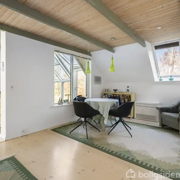 Et rum med et rundt bord dækket med dug, to stole og en grå lænestol. To grønne loftlamper hænger over bordet. Lyst træloft og gulv skaber en hyggelig atmosfære.