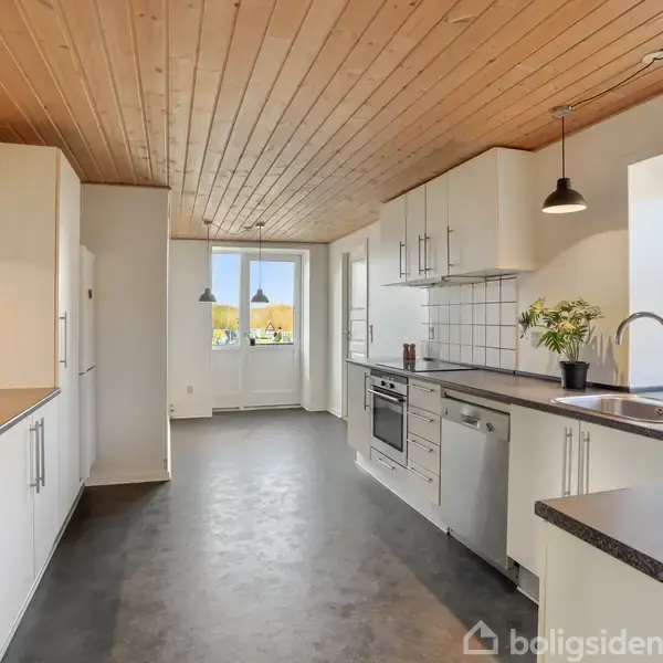 Et moderne køkken med hvide skabe og sorte bordplader, der åbner ud mod en udsigt gennem et vindue. Træloft og hængende lamper skaber en varm atmosfære.