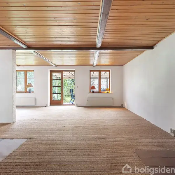 Brændeovn opvarmer et rummeligt værelse med trægulv og træloft. Rummet har store vinduer og en glasdør, der åbner mod en grøn have udendørs.