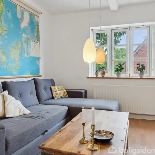 Grå sofa står i en stue med et Danmarkskort på væggen. Træbord med lysestager og skål foran. Tyklagede puder ligger ovenpå, lampe hænger over. Vindue viser bygning udenfor.