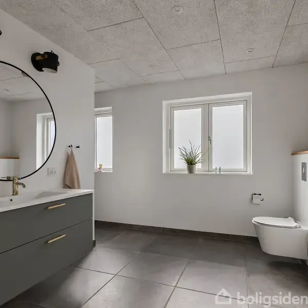 Spejl hænger over en grå vask med skuffer; en plante står på vindueskarmen. Toilettet er væghængt. Rummet har lyse vægge og gulvfliser. Hylder med genstande tilføjer indretningen personlighed.