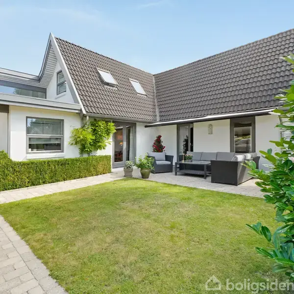 Hus med mørkt tegltag, sidder i en velplejet have med græsplæne og flisebelagt terrasse. Der står havemøbler og planter på terrassen. Skyfri himmel ses i baggrunden.
