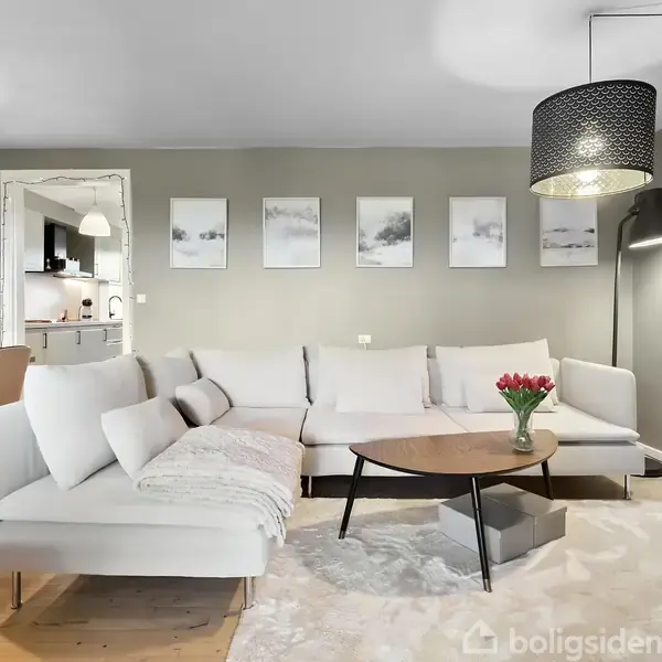 En lys stue med en hvid hjørnesofa står mod en grå væg dekoreret med billeder. Et sofabord holder en vase med røde blomster. Bagved ses et køkken.