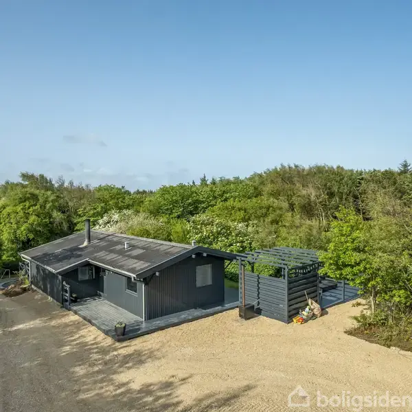 Hus omgivet af grønne træer, står stille på en grusbelagt gårdsplads. Skyfrit blå himmel ses ovenover, med frodig vegetation rundt om huset og indkørsel i forgrunden.