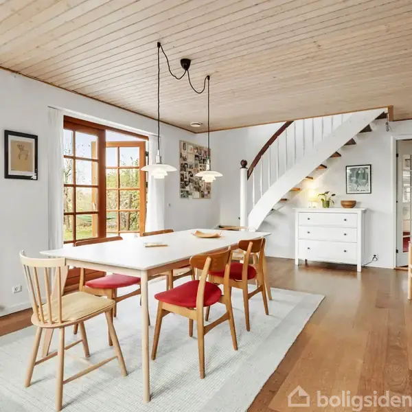 Et lyst spisebord med træstole står i et rum med trægulv og -loft, omgivet af vinduer og en fransk dør. En trappe fører op til en anden etage.