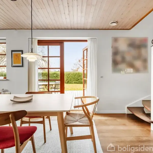 Et spisebord med træstole og røde puder står i et lyst rum. En trappe fører opad til højre, og døre fører ud til en terrasse med havemøbler.