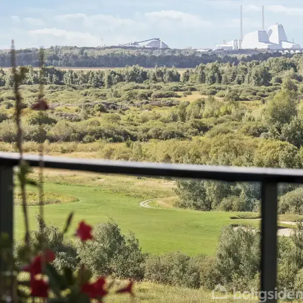 Blomster i en altankasse står stille, hvoralt fra terrassen strækker udsigten sig ud over frodige, grønne marker med træer og en fjern industrihimmel i horisonten.