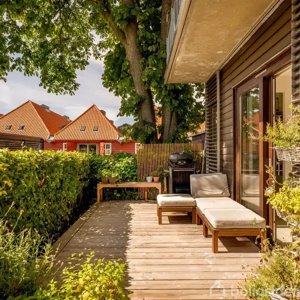 En terrasse med to liggestole og en grill står solbadet under et stort træ. Omgivet af grønne planter og tætklippede hække, ses røde hustage i baggrunden.