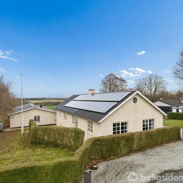 En hvid bungalow med solpaneler på taget omgivet af en velplejet have og indkørsel, beliggende i et landligt område med åbne marker og træer i baggrunden.